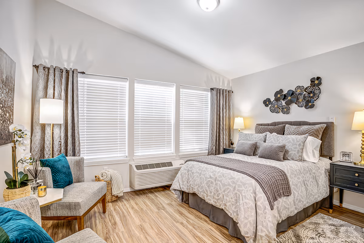 A bright and cozy bedroom with a large bed adorned with multiple pillows and a patterned bedspread. The room features three large windows with blinds and curtains, two bedside tables with lamps, a decorative wall art above the bed, and a seating area with two gray chairs and teal cushions. The floor is wood, and there is a small rug near the bed.