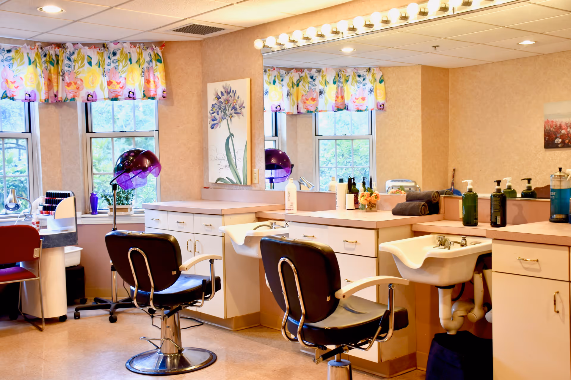 Interior of a salon area in a senior living facility with two black salon chairs in front of sinks and counters. The room has large mirrors, floral curtains on the windows, hair dryers, and various bottles of hair care products on the counters.