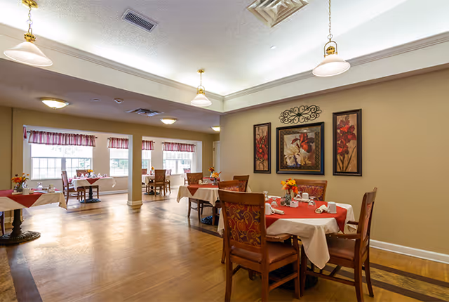 A well-lit dining room with multiple tables covered in white and red tablecloths, each set with napkins and small flower arrangements. The room has wooden flooring, beige walls adorned with framed floral artwork, and several windows with red valances allowing natural light to enter. Ceiling lights hang above the tables providing additional illumination.