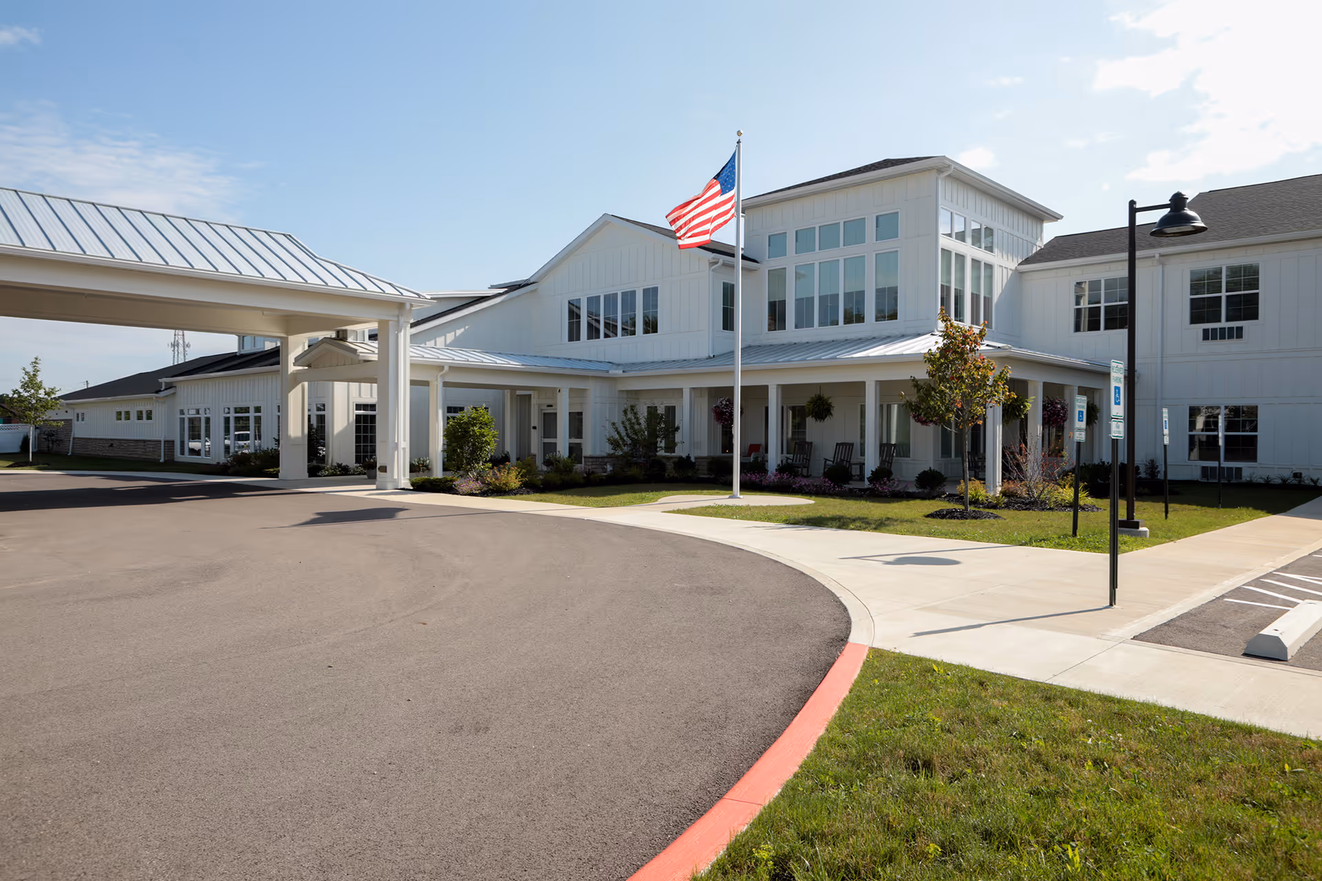 Exterior view of Heritage Crossing senior living facility with white siding, large windows, a covered entrance, an American flag on a flagpole, and a curved driveway with parking spaces and landscaping.