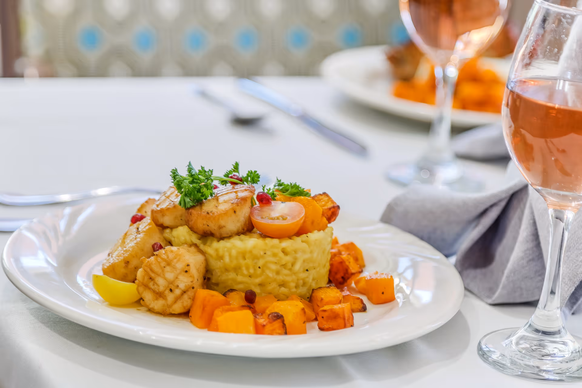A plated gourmet meal featuring seared scallops on a bed of creamy risotto, garnished with fresh parsley and cherry tomato halves, accompanied by roasted cubed vegetables. The plate is set on a white tablecloth with a glass of rosé wine and silverware in the background.