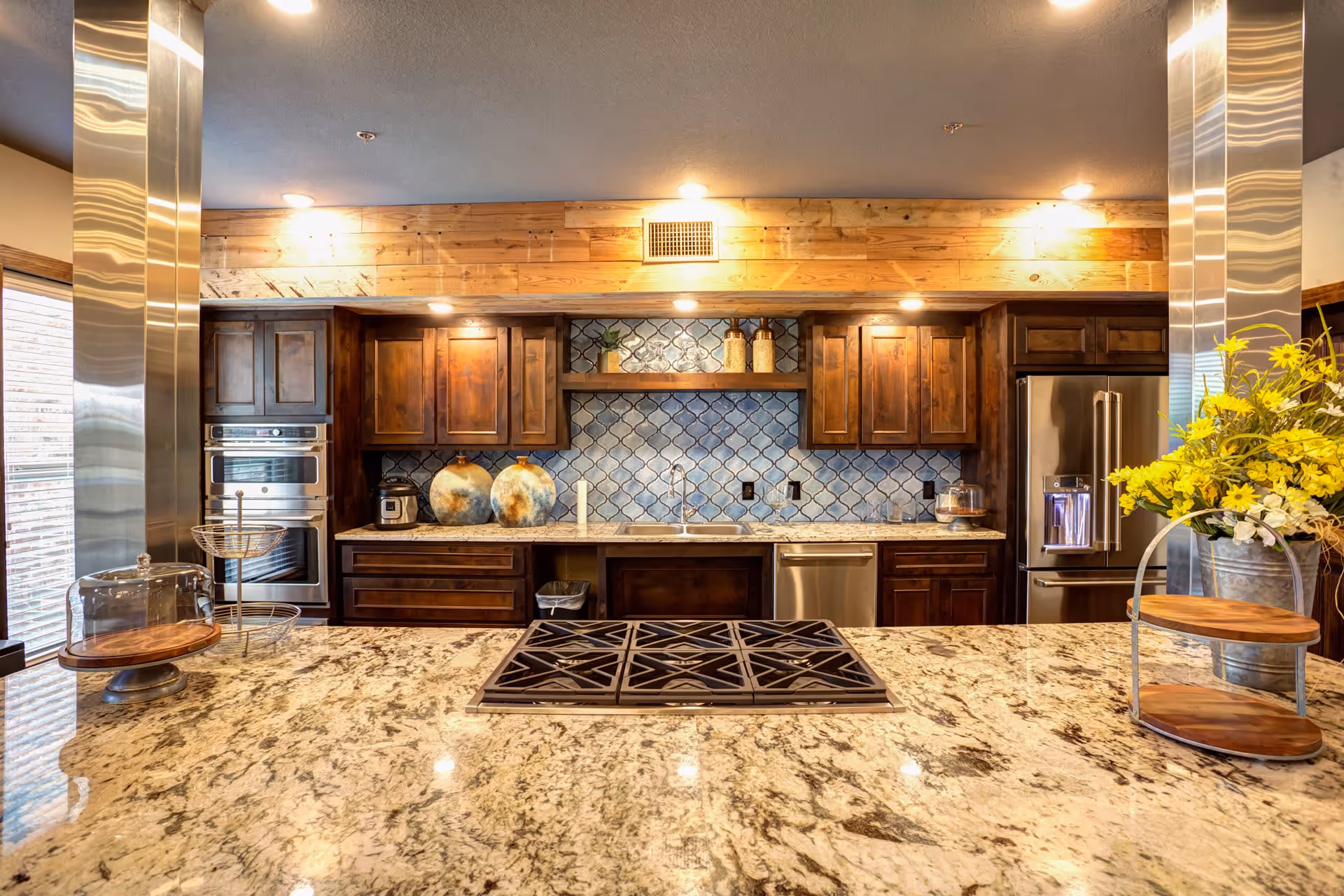 Modern kitchen with granite countertops, a built-in gas stove on the island, dark wooden cabinets, stainless steel appliances including a refrigerator and double oven, decorative blue tile backsplash, and yellow flowers in a metal vase on the right side.