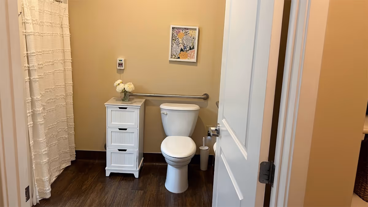 A small bathroom with a white toilet, a white cabinet with three drawers topped with a vase of white flowers, a grab bar on the wall behind the toilet, a floral picture hanging above, and a white shower curtain to the left. The floor is dark wood and the walls are beige.