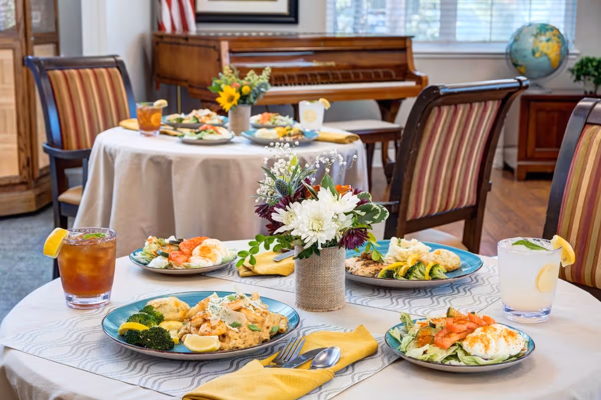 A dining area with round tables covered in white tablecloths, set with plates of food including vegetables, shrimp, and eggs, along with glasses of iced tea and lemonade. A small floral centerpiece is on the table in the foreground. In the background, there is a piano, an American flag, a globe, and striped upholstered chairs around the tables.