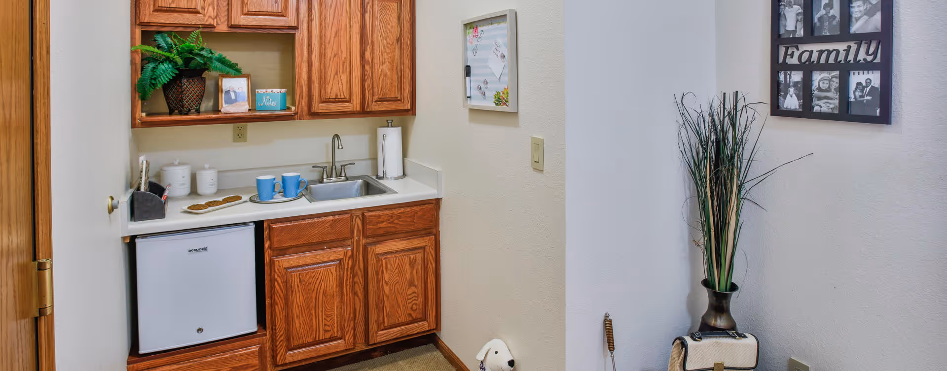 Small kitchenette area with wooden cabinets, a sink, a mini refrigerator, and a countertop holding two blue mugs, a plate with cookies, and some containers. To the right, there is a wall with a family photo collage and a tall vase with decorative grass.