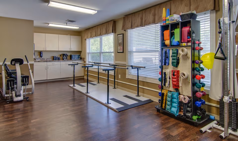 A well-lit rehabilitation or exercise room with wooden flooring, featuring parallel bars for walking exercises, a rack with colorful exercise bands and weights, an exercise bike, and a kitchenette area with cabinets, a microwave, and a coffee maker. Large windows with blinds allow natural light to fill the room.