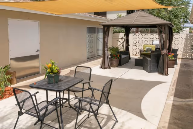Shaded outdoor courtyard with a metal table and four chairs in the foreground and a gazebo seating area with potted plants in the background.