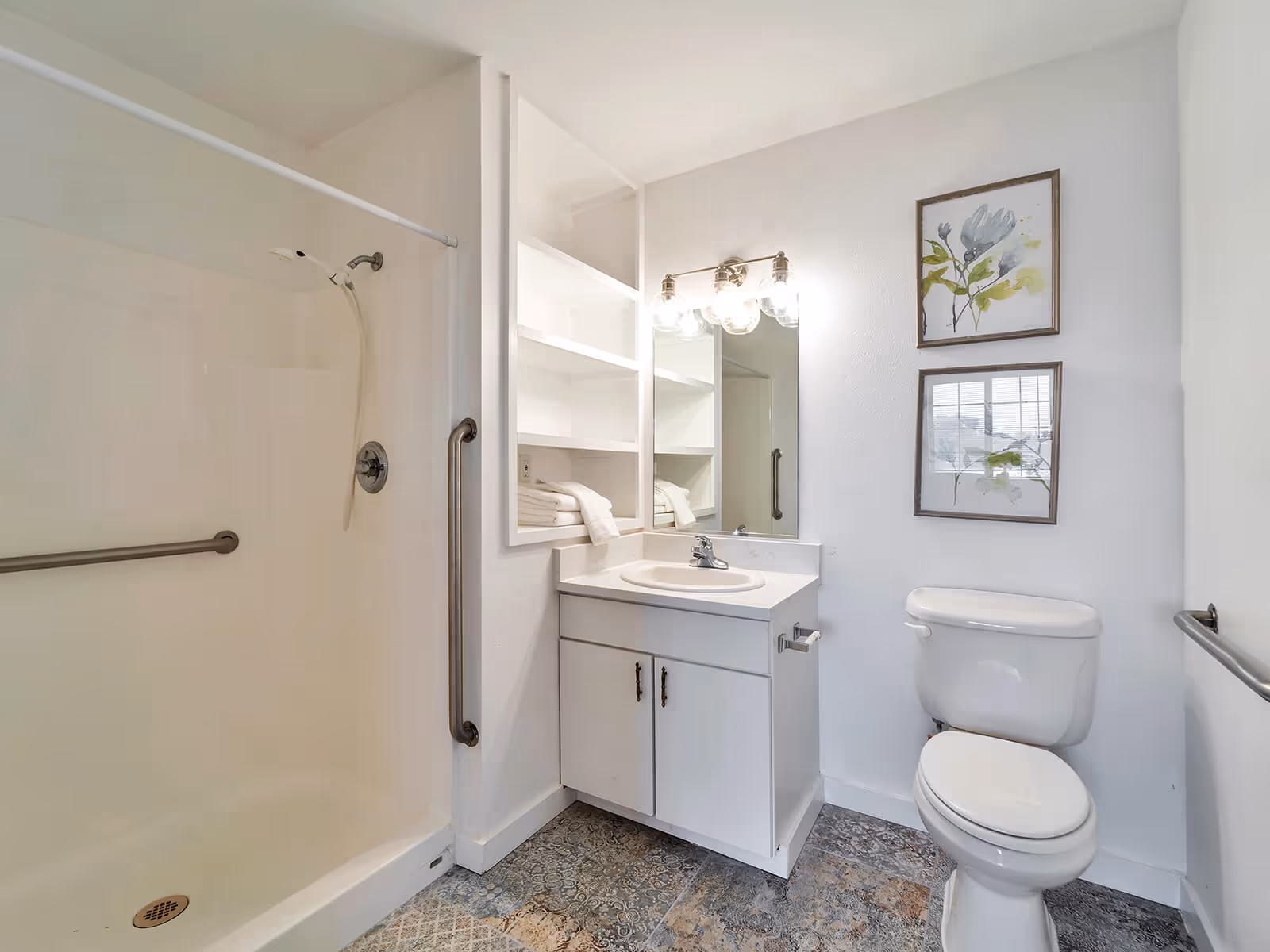 Bright bathroom featuring a walk-in shower with grab bars, a vanity with sink and mirror, open shelving, and a toilet beneath framed artwork.