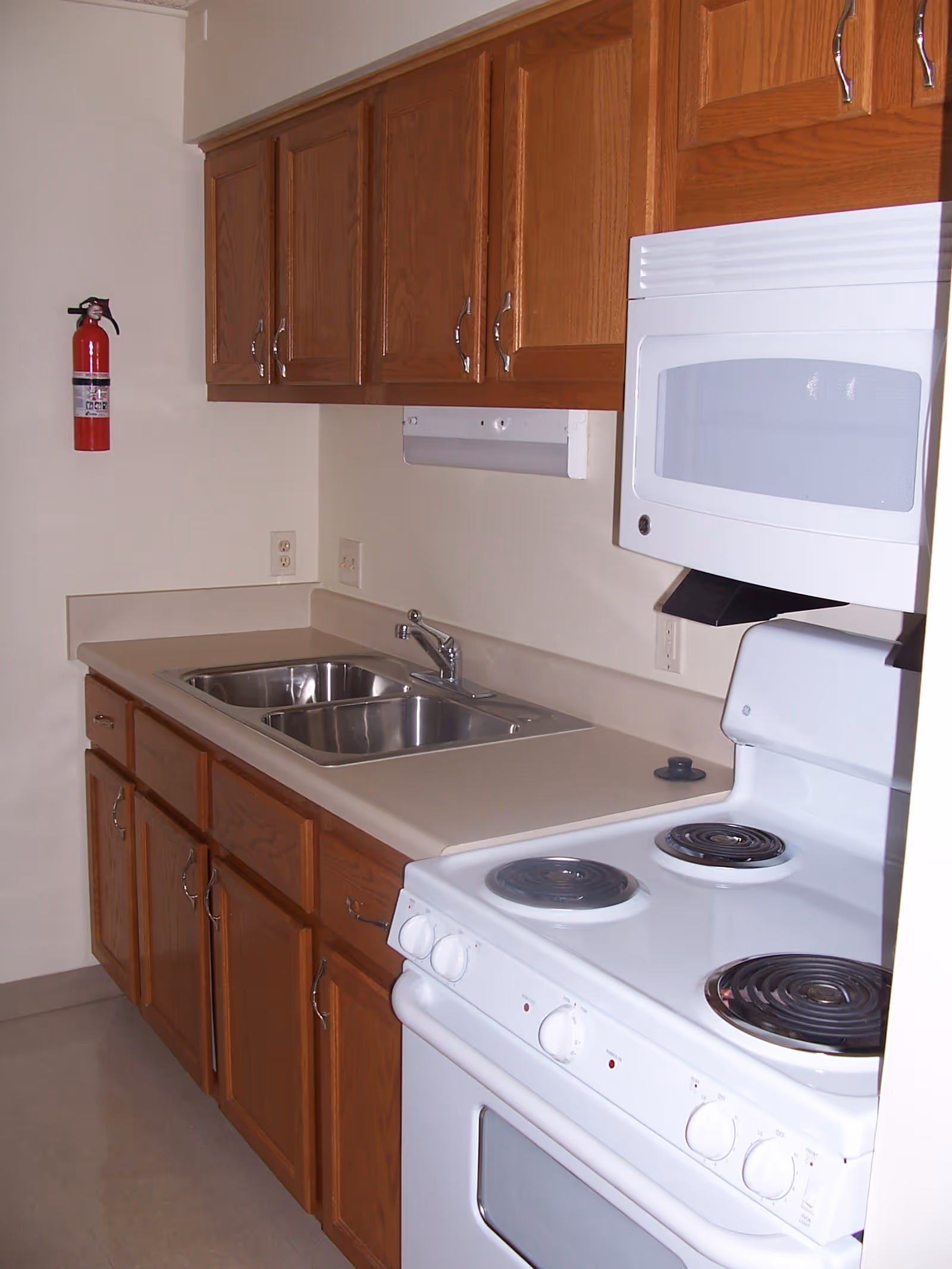 A kitchen area with wooden cabinets, a double stainless steel sink, a white electric stove with coil burners, and a white microwave above the stove. A red fire extinguisher is mounted on the wall to the left.