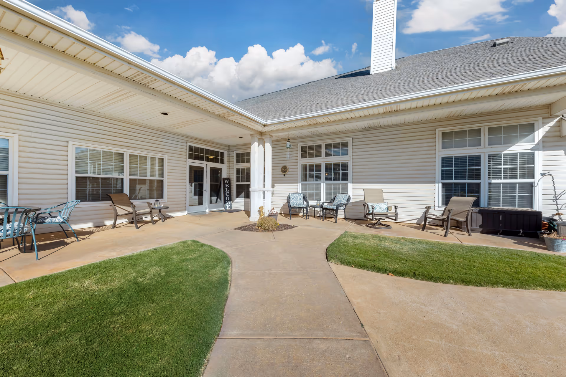 Courtyard patio and entrance of a senior living building with outdoor chairs, walkways, and small lawns under a covered porch.