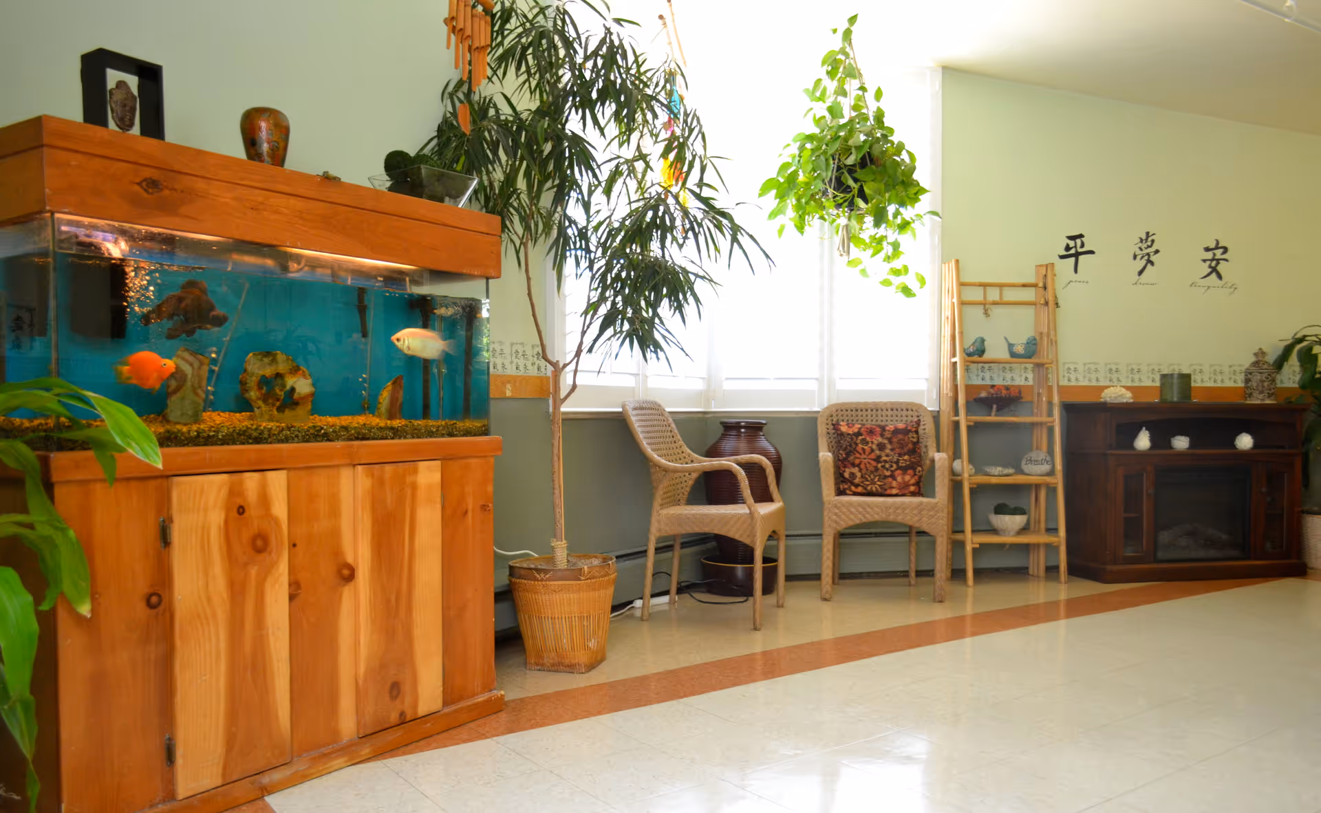 A bright and cozy interior room featuring a large wooden fish tank with colorful fish on the left, two wicker chairs with a floral cushion, several potted plants including a tall one and a hanging one near a window with white blinds. There is a wooden ladder shelf with decorative items and a dark wooden electric fireplace on the right. The wall has Asian characters and a light green and white color scheme.