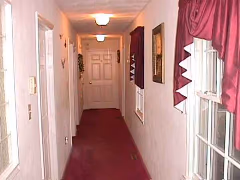 A narrow interior hallway at Jeanne's Elderly Care with windows, burgundy carpet, and pink curtains.
