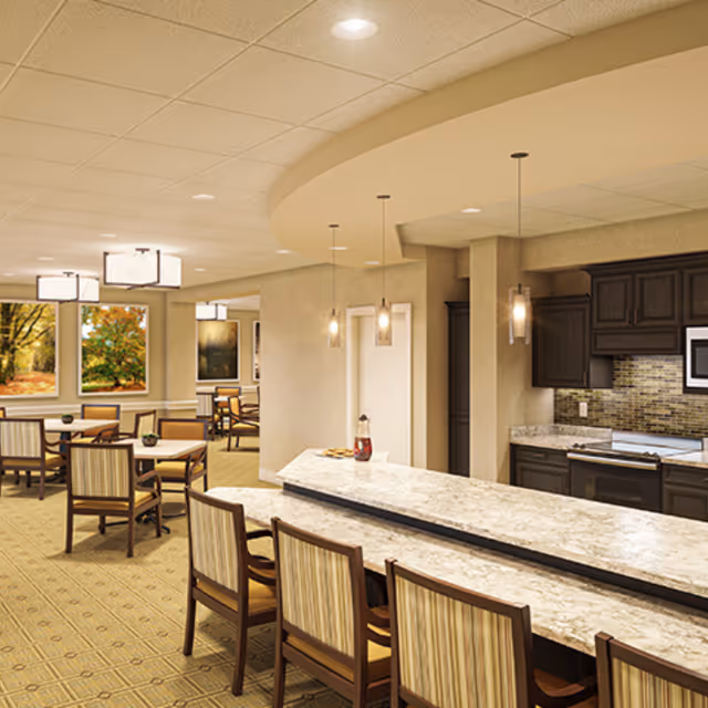 A spacious dining area with multiple tables and chairs, featuring a long marble countertop bar with pendant lights hanging above. The room has beige walls, carpeted floors, and large windows showing autumn trees outside. There is a kitchen area with dark wood cabinets and modern appliances in the background.