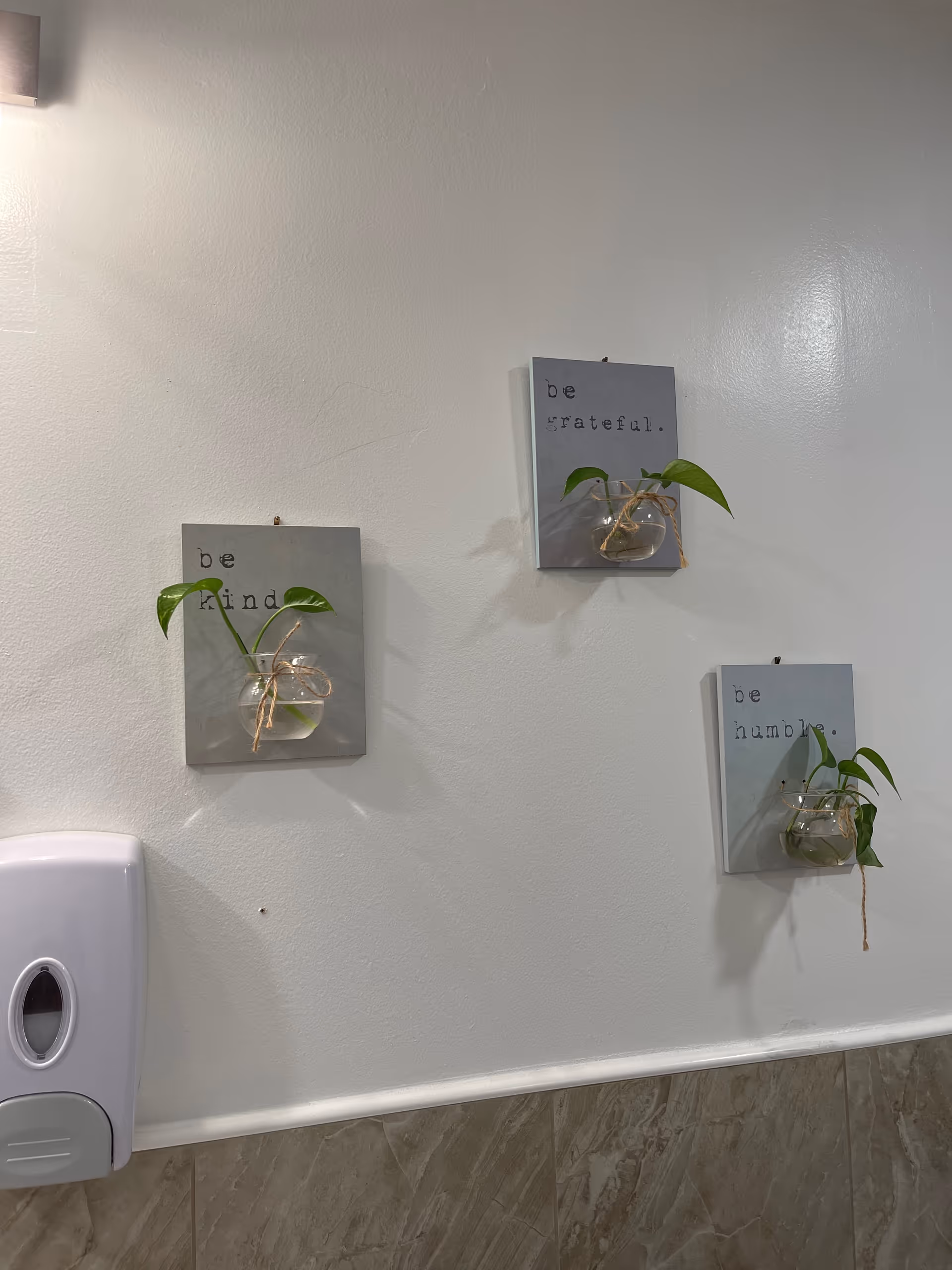 A bathroom wall with three small decorative plaques, each holding a small glass vase with green plant cuttings. The plaques have inspirational words: 'be kind', 'be grateful', and 'be humble'. Below the plaques is a white soap dispenser mounted on the wall, and the lower part of the wall is tiled with beige marble-like tiles.
