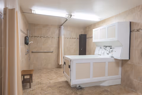 A spacious, tiled shower room with beige walls and floor tiles. The room features a large, white accessible shower tub with controls on the side, a wall-mounted showerhead, a grab bar, a small wooden bench, and a shower curtain for privacy.