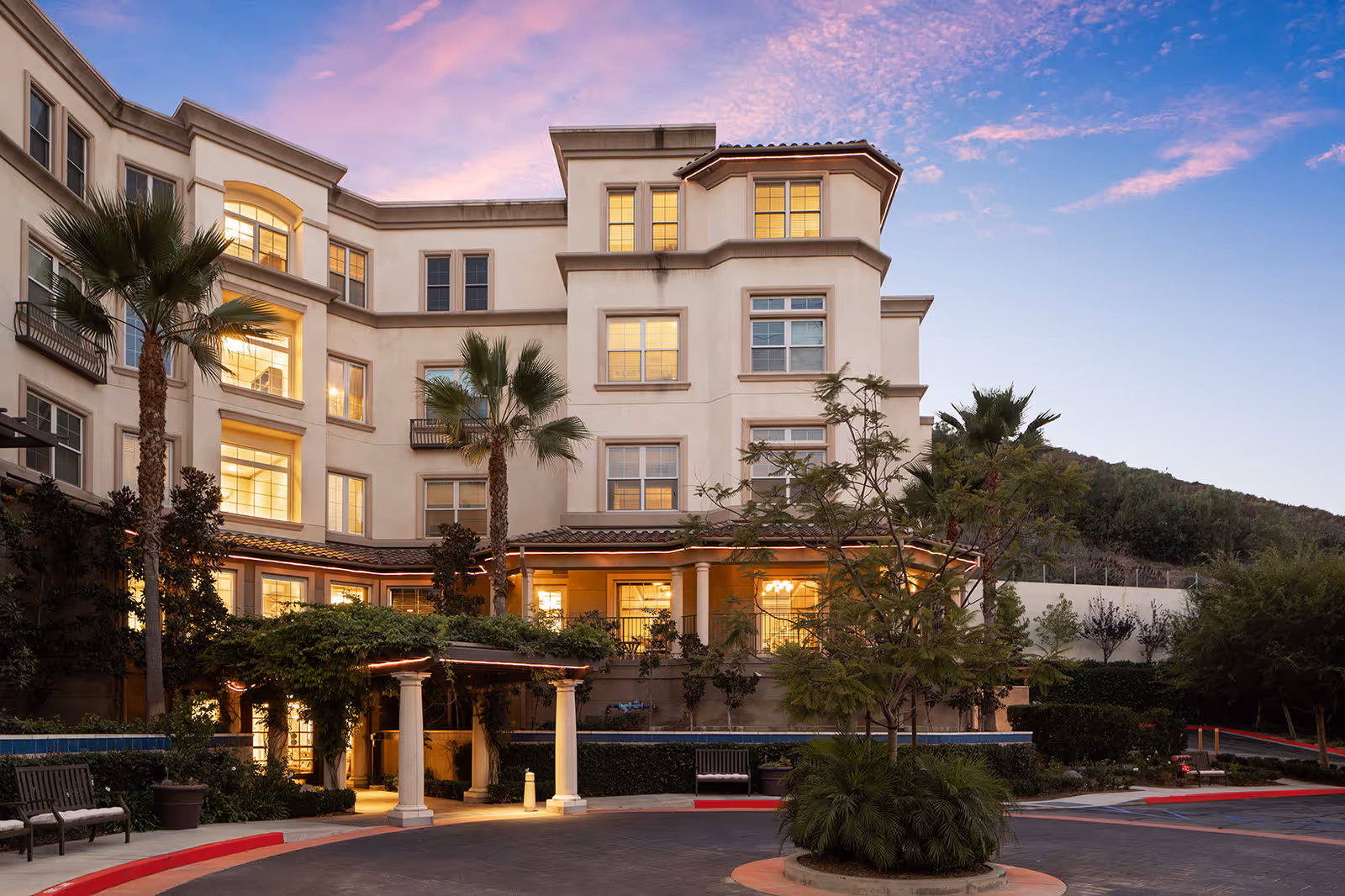 Exterior view of a multi-story senior living facility building at dusk with warm lights glowing from the windows. The building is surrounded by palm trees, shrubs, and a circular driveway with benches and a covered entrance. The sky is partly cloudy with a pink and blue sunset.