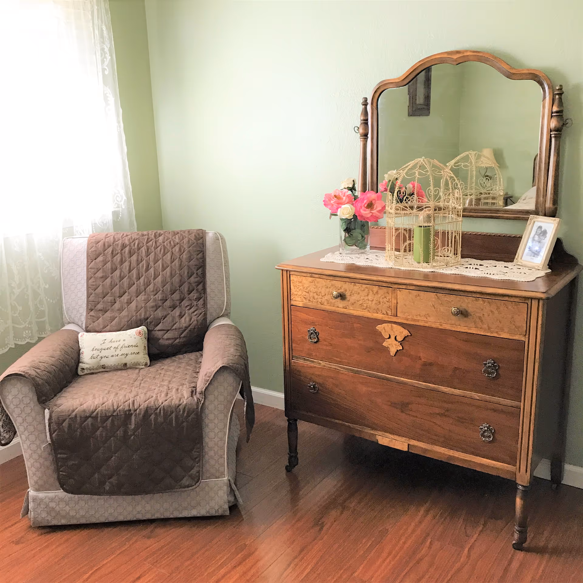 A cozy room corner with an upholstered armchair beside a wooden dresser topped with a mirror, flowers, and decorative accents.