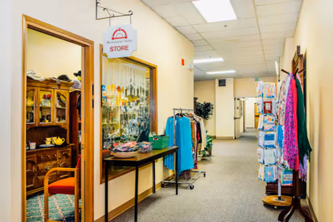 Hallway inside a senior living facility with a small store on the left displaying various items including clothes and accessories. The hallway has racks with clothing and a rotating stand with greeting cards or pamphlets. The walls are light-colored and the ceiling has fluorescent lighting.