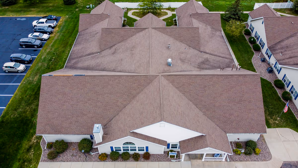 Aerial view of a senior living facility building with a large brown roof, surrounded by green lawns, a parking lot with several cars, and adjacent buildings. The facility has a white exterior with blue window shutters and landscaped bushes around the perimeter.