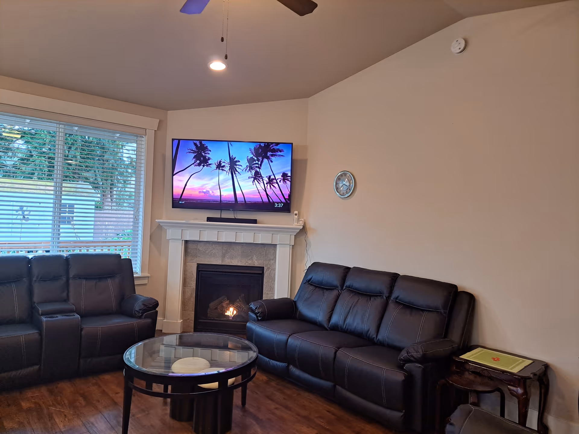 Living room with black leather sofas, a glass coffee table, fireplace and wall-mounted TV displaying a palm tree scene.