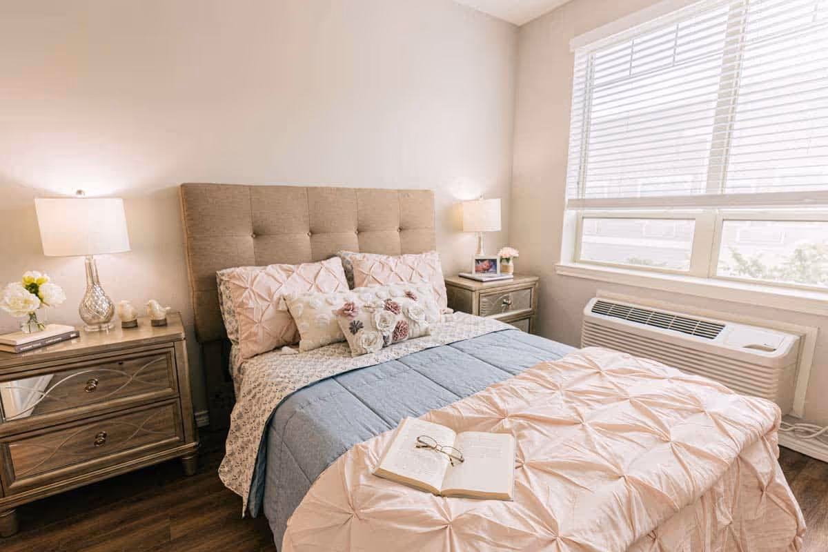 A cozy bedroom with a beige upholstered headboard, a bed made with a light pink and blue comforter, decorative pillows, and an open book with reading glasses on the bed. There are two matching wooden nightstands on either side of the bed, each with a lamp and decorative items. A large window with white blinds lets in natural light, and a heating/cooling unit is installed below the window.
