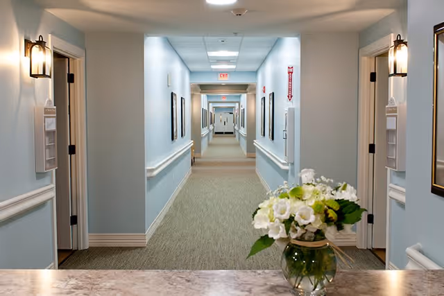 A long, well-lit hallway in a senior living facility with light blue walls, beige carpet, handrails on both sides, and several doors leading to rooms. There are framed pictures on the walls and a vase with white and green flowers on a marble countertop in the foreground.