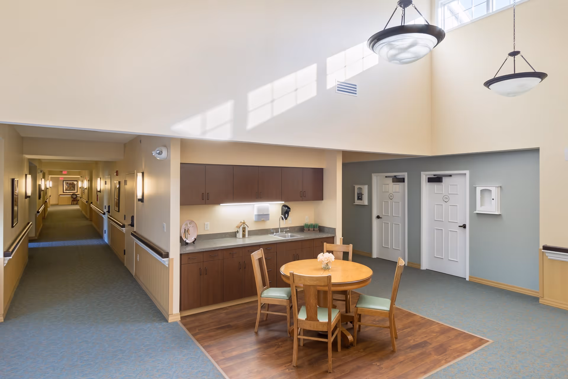 A well-lit common area in a senior living facility featuring a round wooden table with four chairs on a wood-floored section. Behind the table is a countertop with a sink, cabinets, and decorative items. To the right, there are two white doors with room numbers, and to the left, a long hallway with wall-mounted lights and framed pictures. The ceiling is high with two hanging light fixtures and windows allowing natural light to cast shadows on the wall.
