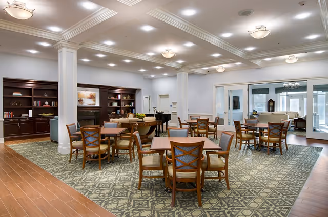 A spacious, well-lit common area with multiple wooden tables and chairs arranged on a patterned carpet. The room features white walls, decorative ceiling lights, and large white columns. In the background, there is a built-in dark wood bookshelf with decorative items and a fireplace. A grand piano is also visible near the center of the room. Beyond the main area, there is a seating area with sofas and large windows allowing natural light to enter.