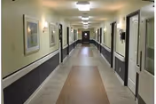 A long, well-lit hallway in a senior living facility with doors on both sides, handrails along the walls, framed artwork, and ceiling lights evenly spaced.