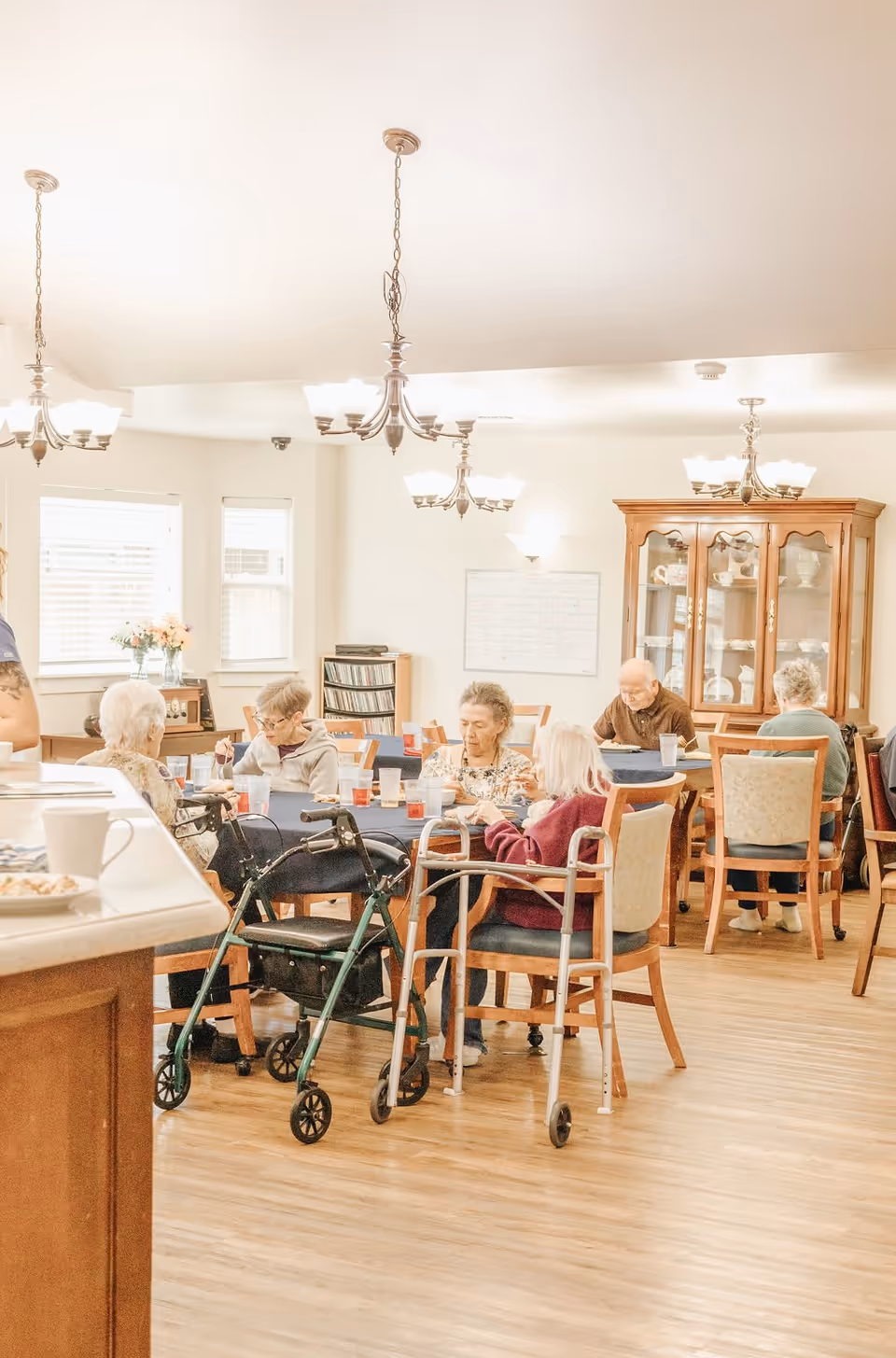 A group of elderly people sitting around tables in a well-lit dining area, eating and socializing. The room has wooden floors, chandeliers, and a wooden cabinet displaying dishes. Walkers are positioned near some of the chairs.