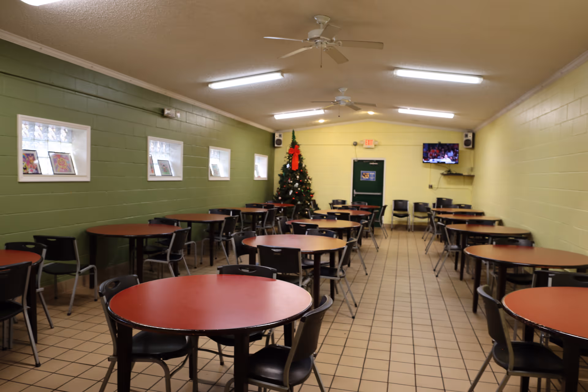 Large dining/activity room with multiple round tables and chairs, green walls, a Christmas tree at the far end and a wall-mounted TV.