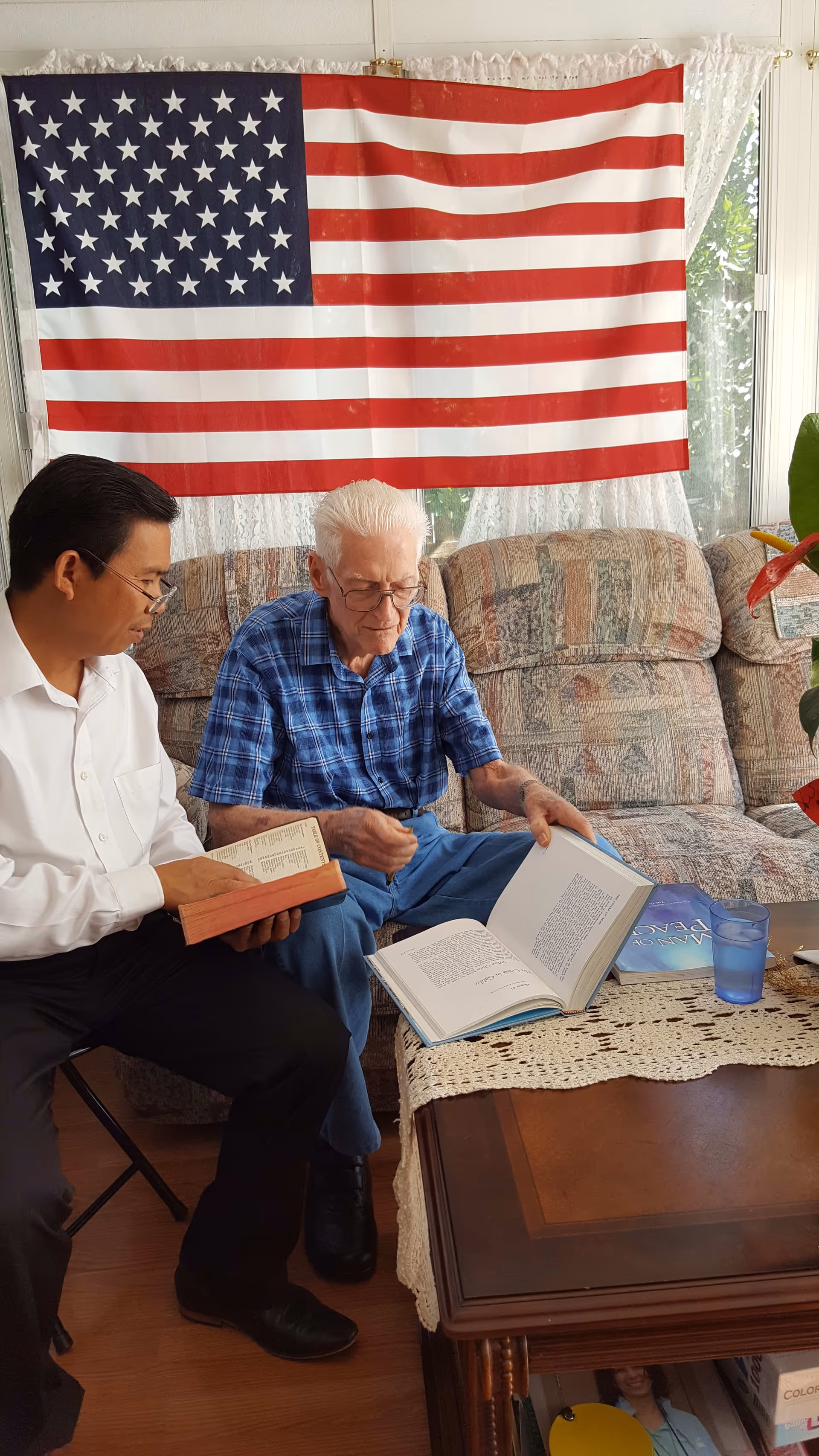Two men sitting in a living room with an American flag hanging on the wall behind them. One man, wearing a blue plaid shirt and glasses, is looking at an open book on a wooden coffee table with a lace runner. The other man, dressed in a white shirt and black pants, is holding a book and looking at it. The room has a patterned couch and a window with lace curtains.