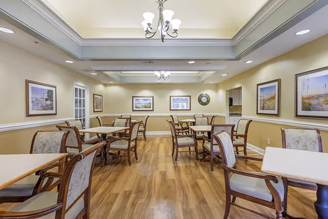 Bright dining room with wooden floors, multiple tables and upholstered chairs under coffered ceilings and chandeliers.