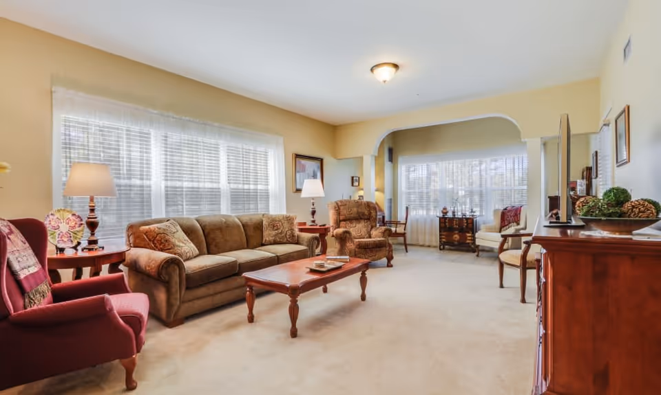 A spacious living room with beige walls and carpet, featuring a brown sofa, two armchairs, a wooden coffee table, side tables with lamps, and large windows with white blinds and sheer curtains allowing natural light to fill the room.