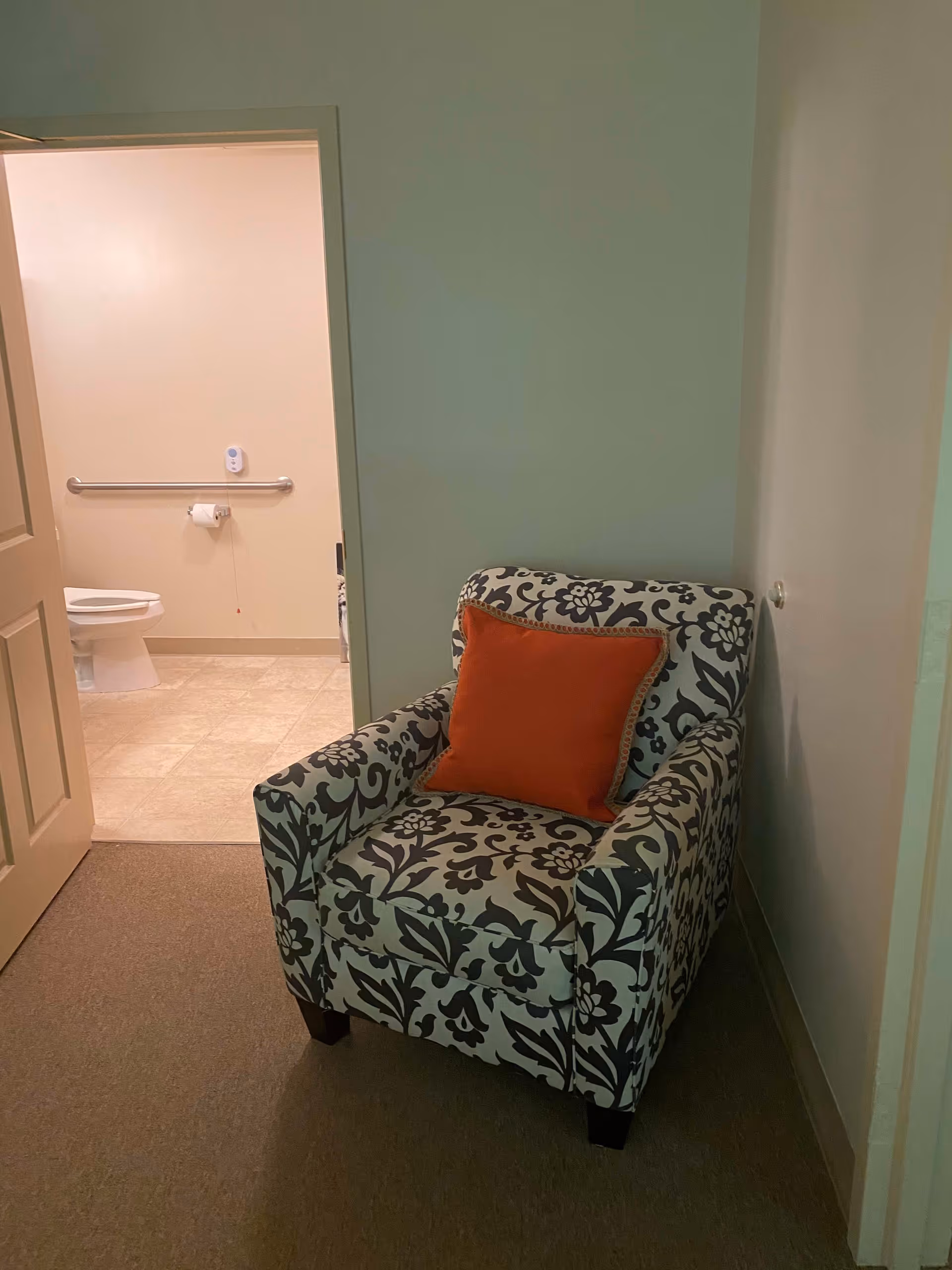 A patterned armchair with a red cushion is placed in a corner of a room with beige walls and carpeted floor. An open door reveals a bathroom with a toilet, grab bar, and tiled floor.