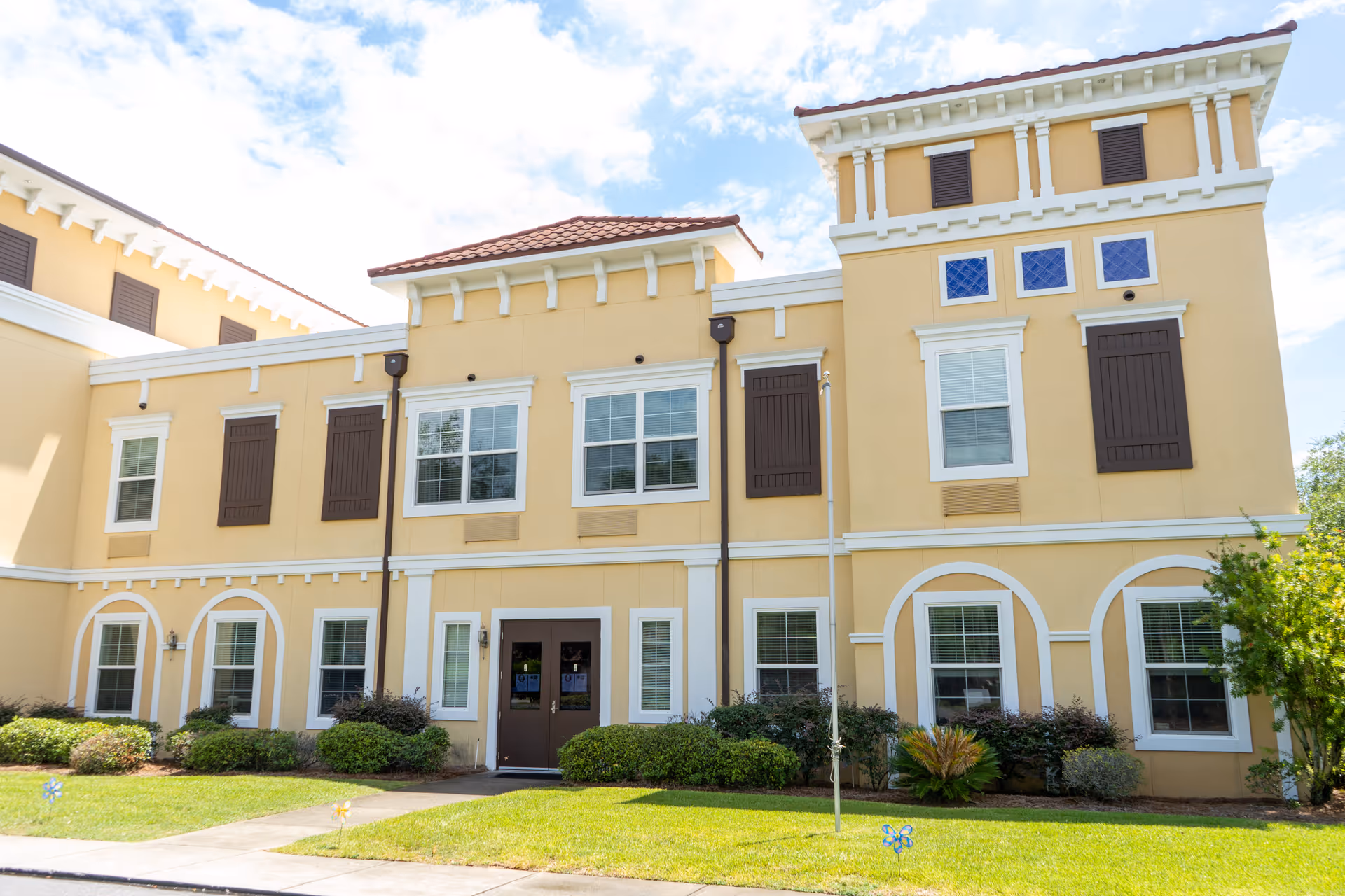 Exterior view of a two-story building with beige walls, white trim, and brown shutters. The building has multiple windows, a double door entrance, and a well-maintained lawn with shrubs and small plants in front. The sky is partly cloudy.