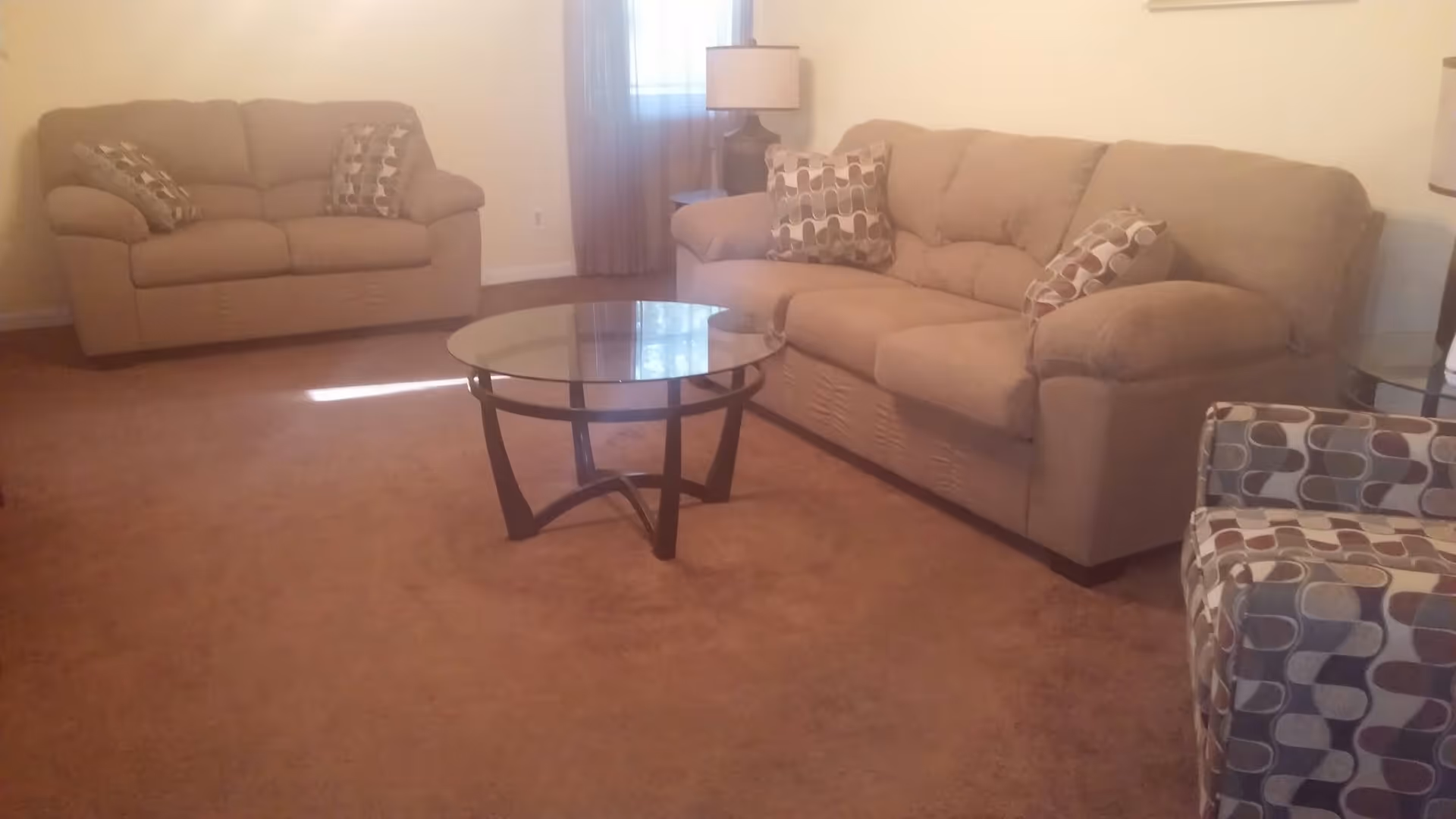 A living room with two beige sofas, a patterned armchair, a round glass coffee table, and a floor lamp on brown carpet.