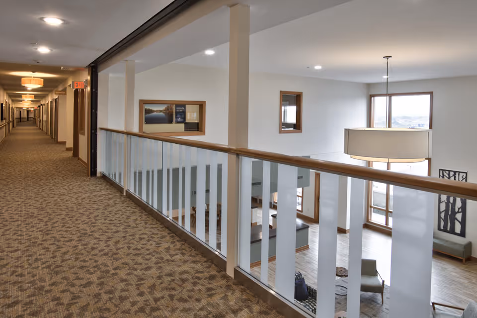 Carpeted second-floor hallway with a wood-and-glass railing overlooking a two-story lobby with large windows and hanging light fixtures.