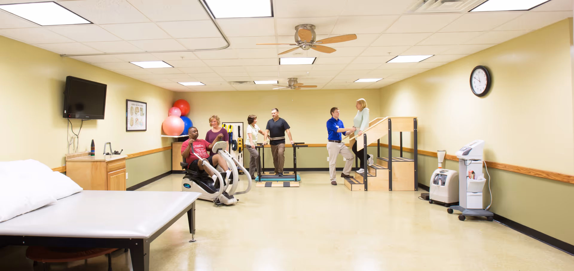 A physical therapy room with exercise equipment and therapy tools. Five people are present: one person is using a recumbent exercise bike, two people are standing on a balance platform with a therapist, and another therapist is assisting a woman walking up wooden stairs with handrails. The room has beige walls, ceiling fans, a wall clock, and a mounted TV.