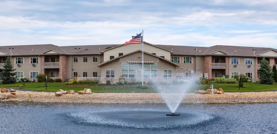 Front exterior of a two-story senior living building with a central flagpole and a fountain in a pond in the foreground.