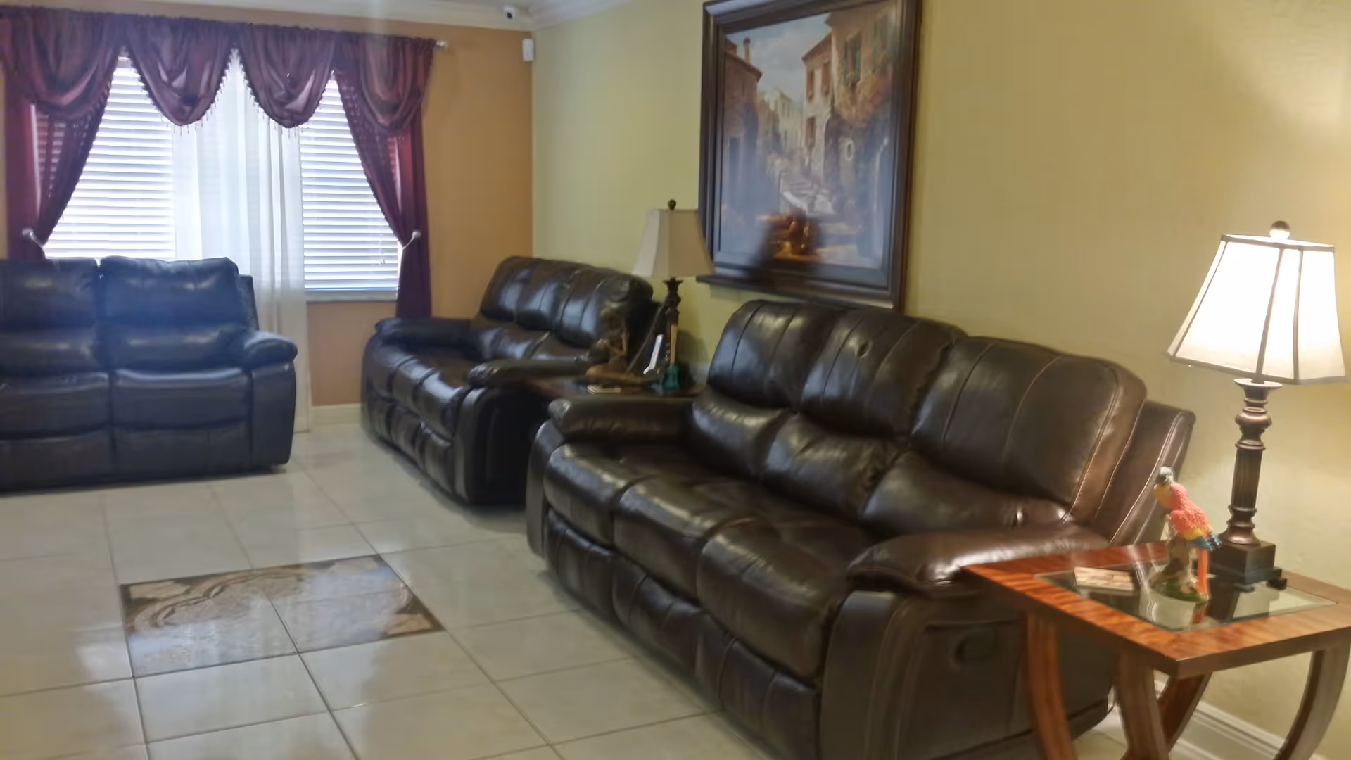 A living room with two dark brown leather sofas, a wooden side table with a lamp and a colorful parrot figurine, a second lamp on a small table between the sofas, a large framed painting on the wall, and a window with white blinds and dark red curtains.
