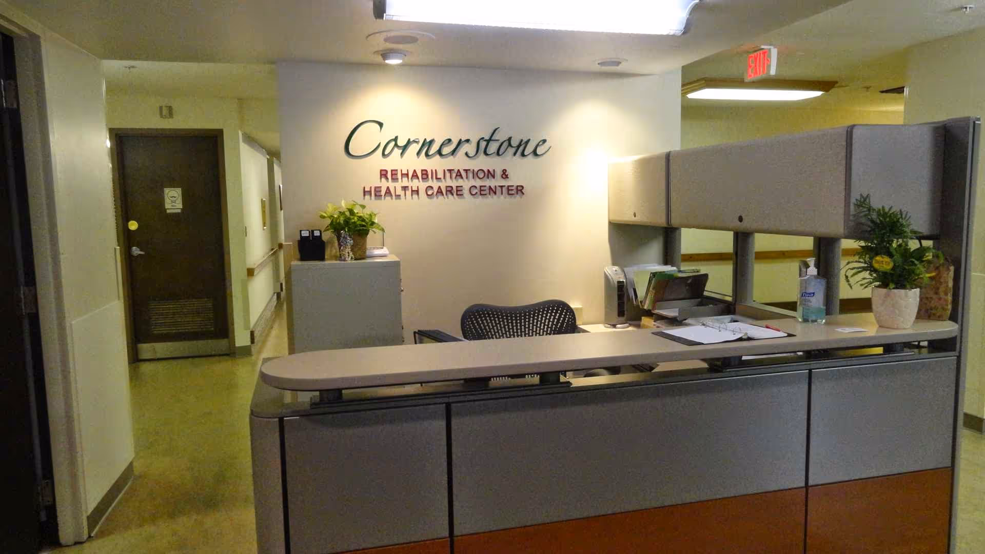 Reception desk area inside Cornerstone Rehabilitation & Health Care Center with a chair, office supplies, plants, and a sign on the wall behind the desk.