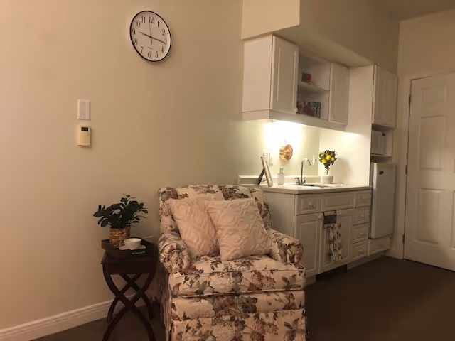 Floral armchair and side table in a small living area beside a compact kitchenette with white cabinets and a wall clock.