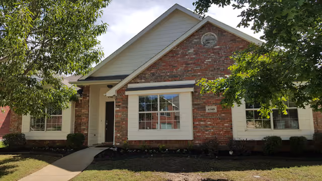 Front exterior view of a single-story brick residential building with white trim, large windows, and a small covered entrance. There are trees and shrubs in the front yard and a concrete walkway leading to the door.