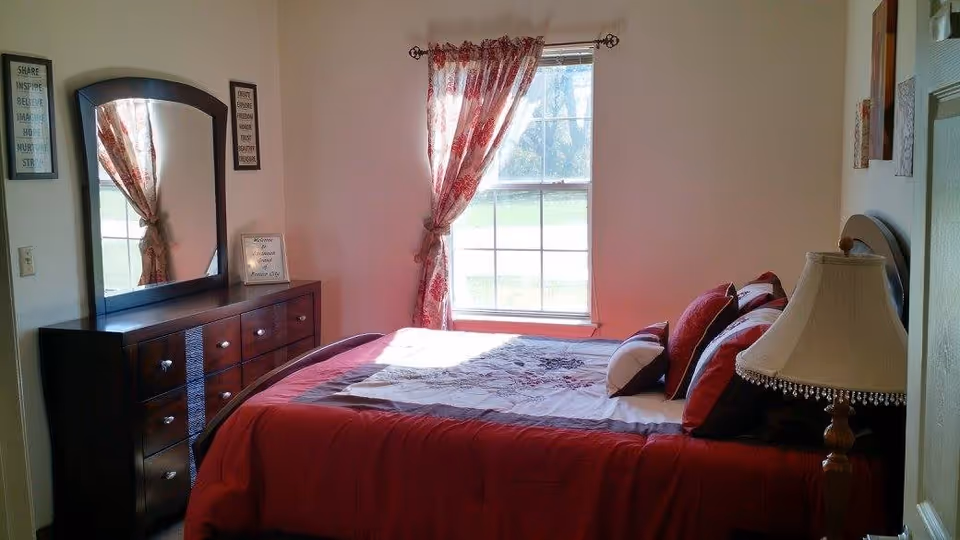 A bedroom with a large window covered by red and white patterned curtains. The bed has a red bedspread with multiple pillows in red, white, and brown. There is a wooden dresser with a large mirror on the left side of the room, and a lamp with a beige lampshade on a nightstand to the right of the bed. The walls are light-colored with framed artwork.