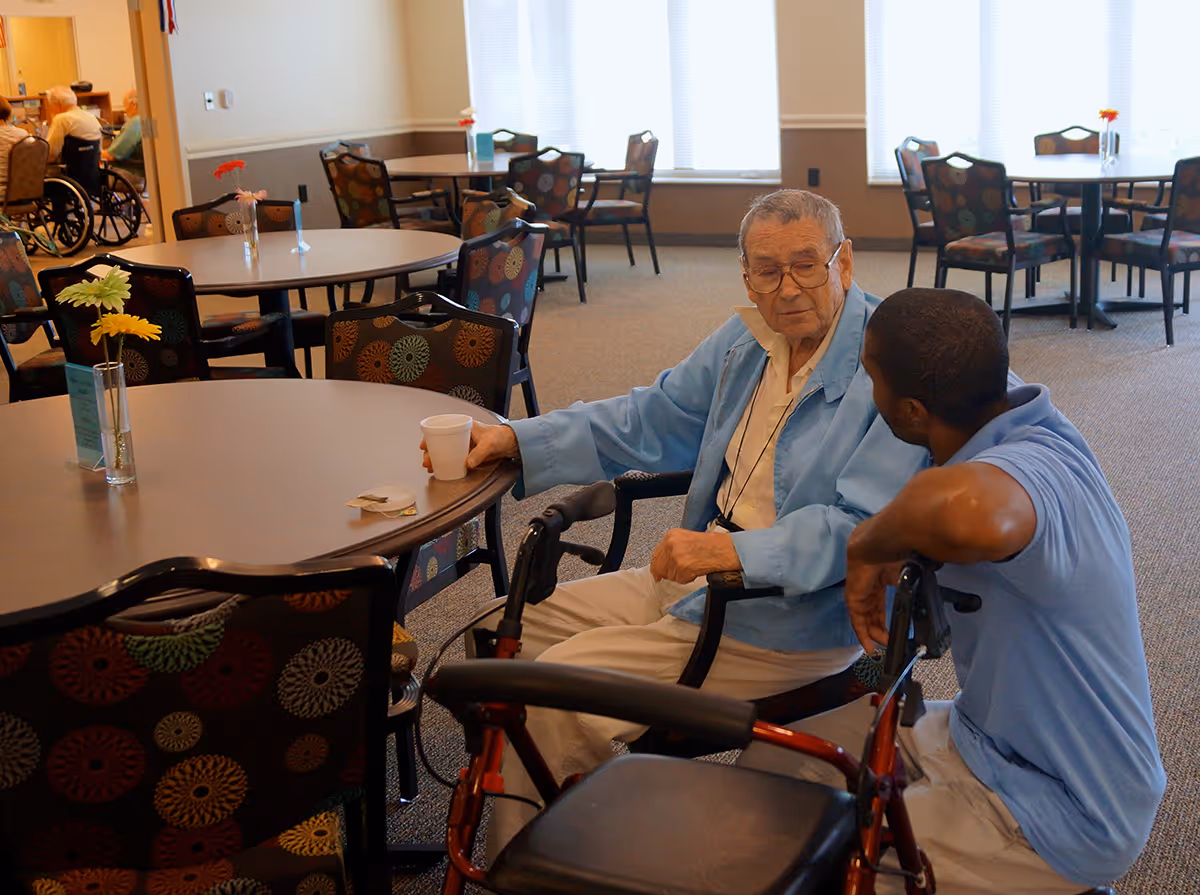 An elderly man in a light blue jacket sitting at a round table with a cup in his hand, engaged in conversation with a younger man in a blue shirt. The room has multiple round tables with patterned chairs and small vases with flowers, and other elderly people are visible in the background.