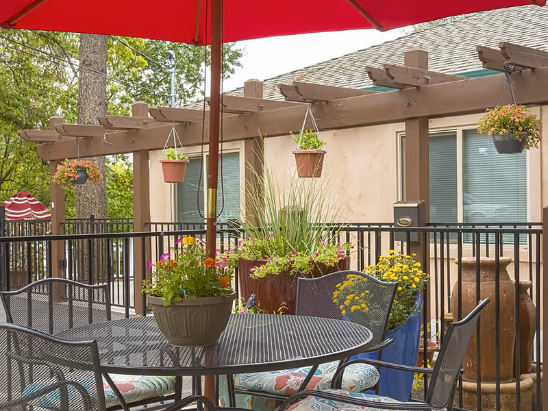 Outdoor patio area with a round metal table and four chairs with floral cushions. A large red umbrella provides shade. Several hanging flower pots and large planters with colorful flowers and greenery surround the space. A beige building with windows and a wooden pergola is in the background.