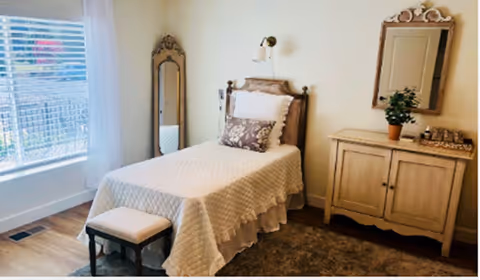 A cozy bedroom with a single bed covered in a beige quilt and decorative pillows. Next to the bed is a small cushioned stool. A tall ornate mirror stands against the wall near a window with white blinds and sheer curtains. On the opposite wall, there is a wooden cabinet with a decorative mirror above it and a potted plant on top.