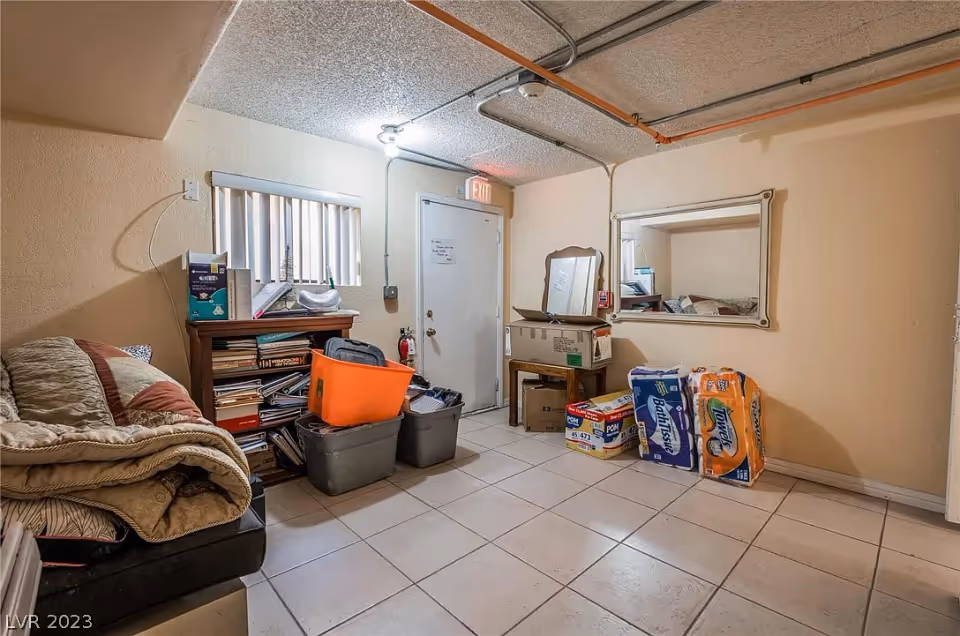 A small room with tiled floor and beige walls, containing a couch with folded blankets, a wooden bookshelf filled with books and various items, several plastic storage bins, a small table with a mirror above it, and packages of paper towels and bath tissue stacked against the wall. There is a door with an exit sign above it and a small window with vertical blinds.
