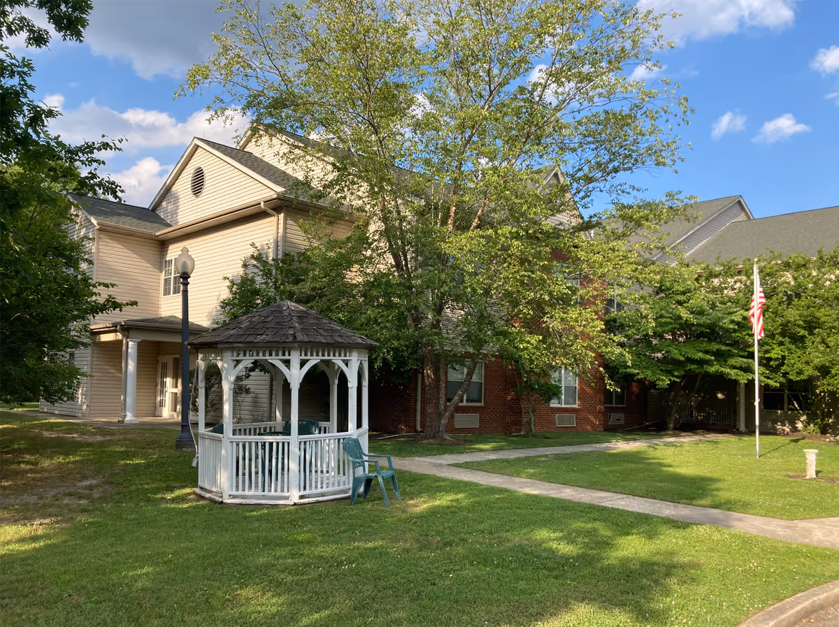 Outdoor view of Woodview Estates showing a two-story building partially covered by trees, a white gazebo with a shingled roof on a grassy lawn, a couple of green plastic chairs next to the gazebo, a sidewalk, a lamppost, and an American flag on a flagpole under a partly cloudy blue sky.
